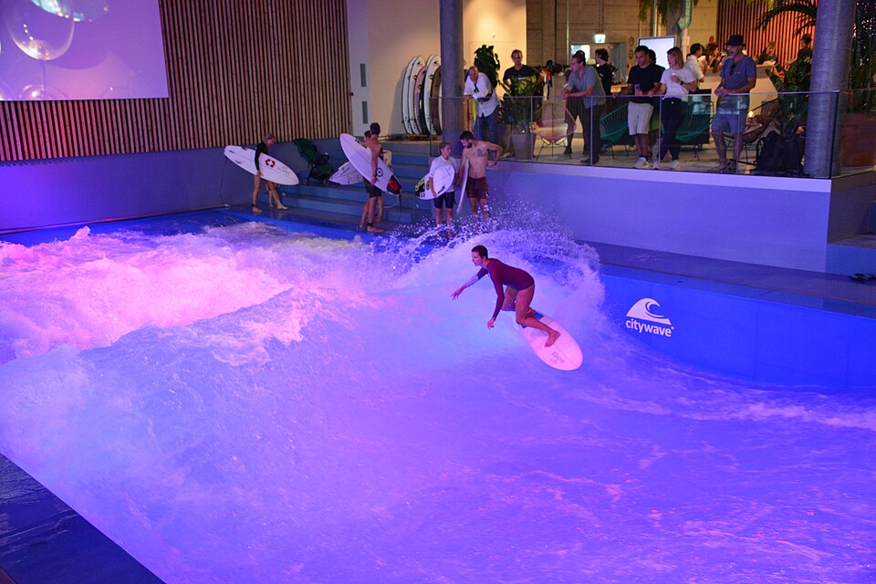Surfer auf einer stehenden Indoor-Welle in der Schweiz, weitere Surfer warten mit Brettern am Beckenrand